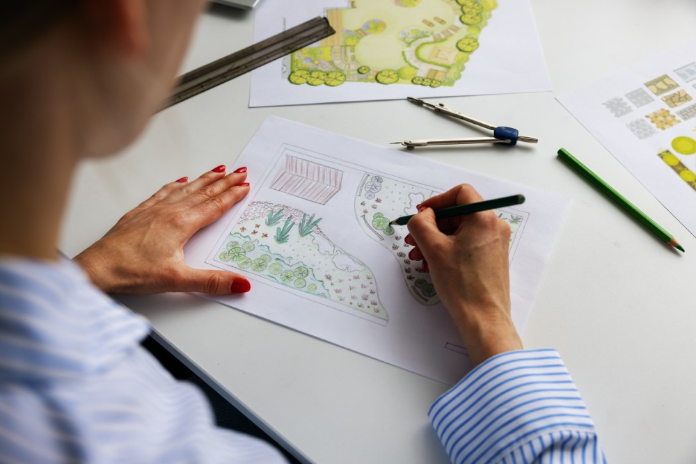 Eine Person zeichnet mit einem Buntstift einen Gartenplan auf einem weißen Schreibtisch.